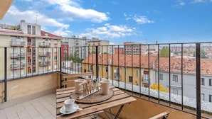 Ein Balkon mit einem Tisch für zwei Personen, mit Blick auf eine Stadtlandschaft mit Gebäuden und blauem Himmel.