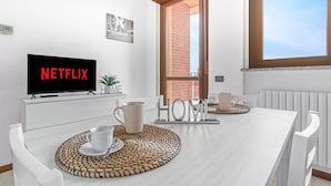 Ein modernes Wohnzimmer mit einem Flachbildfernseher, der 'NETFLIX' anzeigt, einem weißen Esstisch mit zwei Stühlen, einem Couchtisch mit Untersetzer und Blick auf ein Ziegelgebäude durch eine offene Tür.