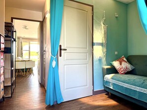 Ein Zimmer mit geschlossener Tür, einem türkisfarbenen Vorhang, einer weißen Wand mit einem Traumfänger, einem türkisfarbenen Sofa mit gemusterten Kissen und einem Holzboden.