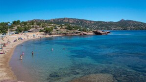 Una playa con agua cristalina, gente nadando y tomando el sol, y un paisaje montañoso al fondo.