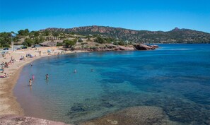 Ein Strand mit klarem Wasser, Menschen beim Schwimmen und Sonnenbaden, und eine hügelige Landschaft im Hintergrund.