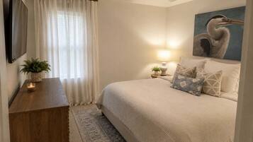 Un dormitorio con cama, mesita de noche, lámpara, cuadro, ventana con cortinas y ventilador de techo.