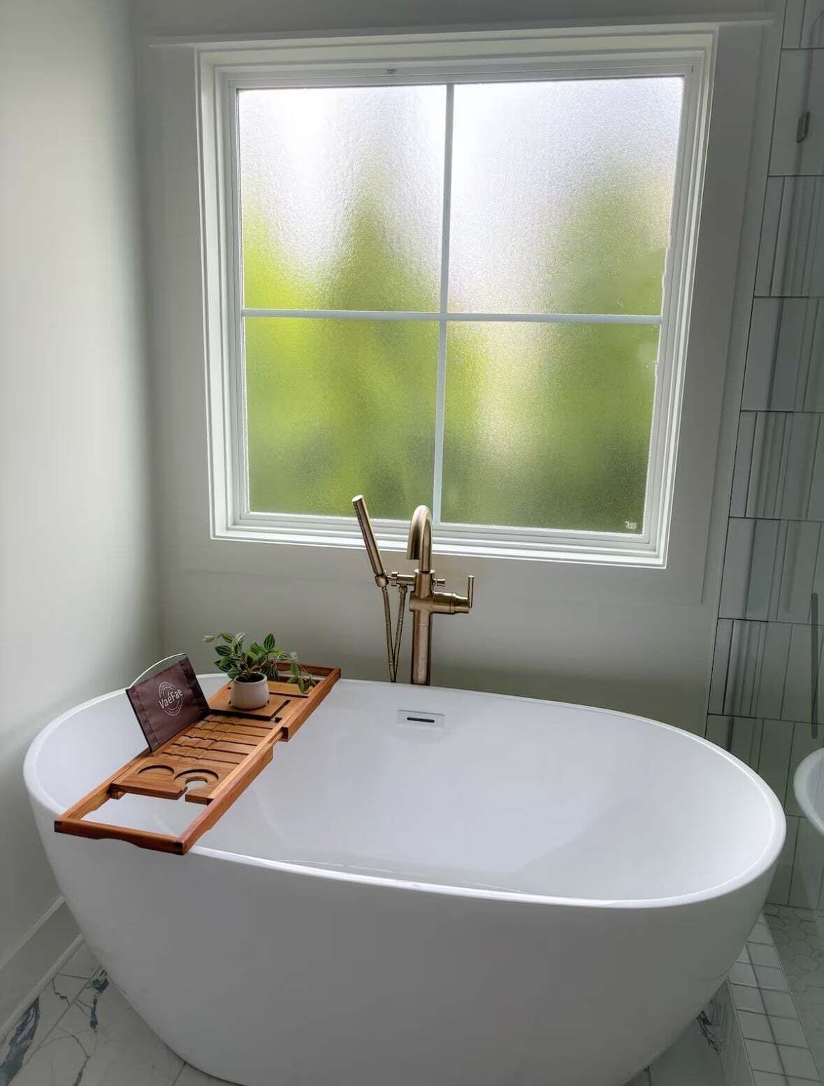 Ein Badezimmer mit freistehender Badewanne, einem Fenster mit Frostglas und einer kleinen Holzablage mit Pflanze und Buch.