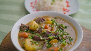 A bowl of stew with meat and vegetables, and a plate of rice in the background. - Vogue Hotel (Karu)