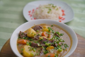 A bowl of stew with meat and vegetables, and a plate of rice in the background. - Vogue Hotel (Karu)
