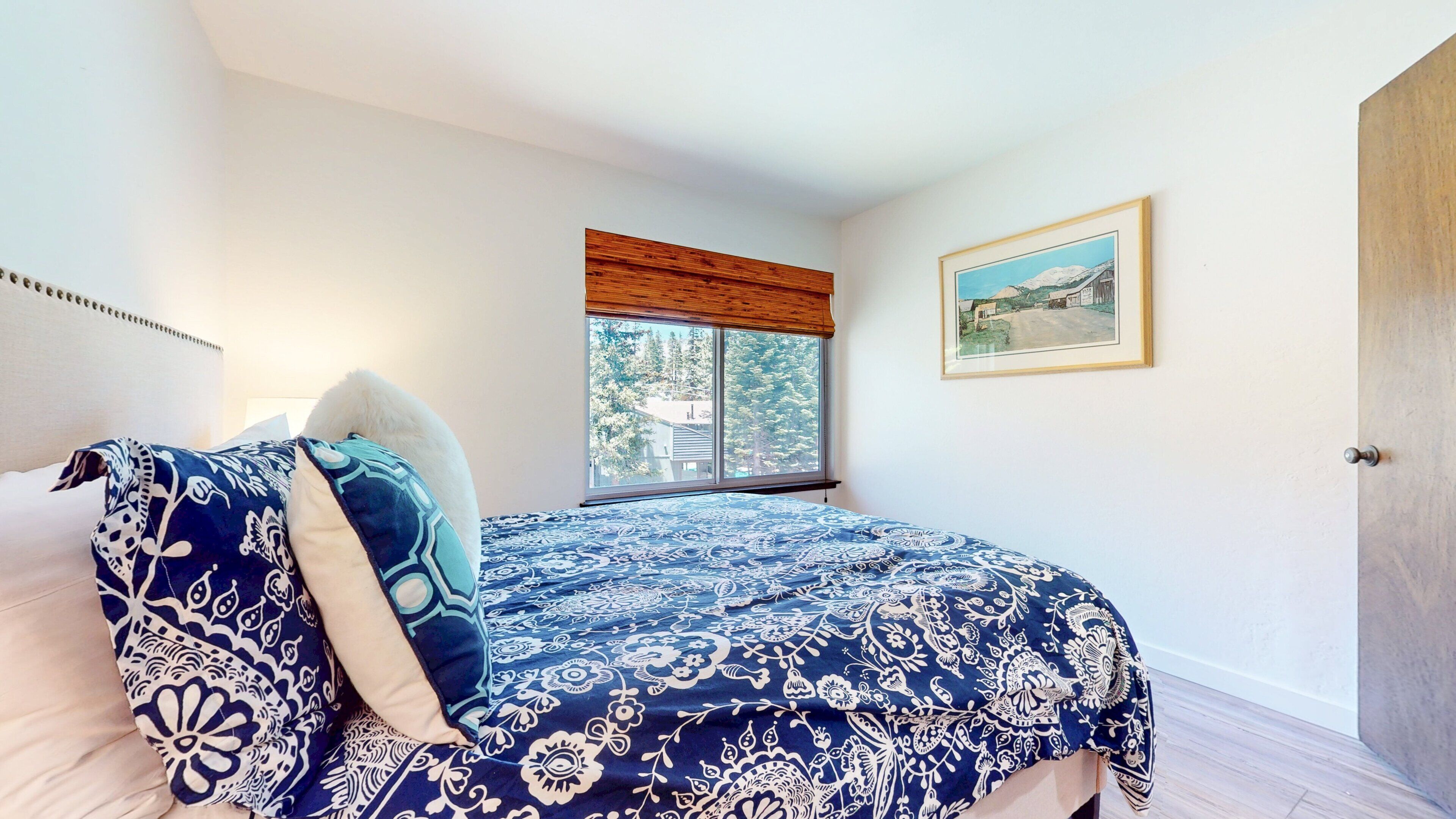 A bedroom with a large bed, a window with a view, a framed picture on the wall, and a door.