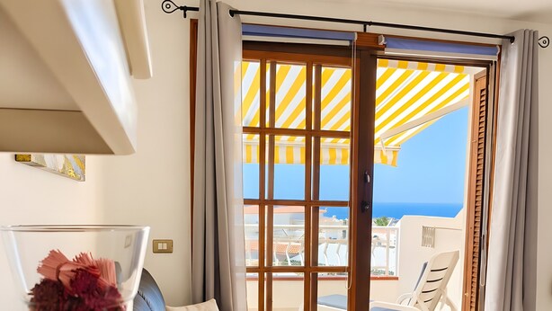 Una habitación con sofá, una mesita de cristal con un jarrón de flores y vista al mar a través de un ventanal con toldo a rayas.