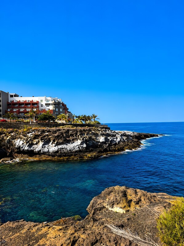 Un hôtel situé sur la côte, avec plusieurs étages, un terrain rocheux et une eau d’un bleu cristallin.