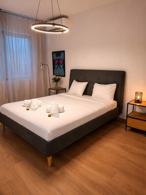 Une chambre d’hôtel moderne avec un grand lit, une table de chevet avec une lampe, une œuvre d’art accrochée au mur et une fenêtre avec des rideaux légers.