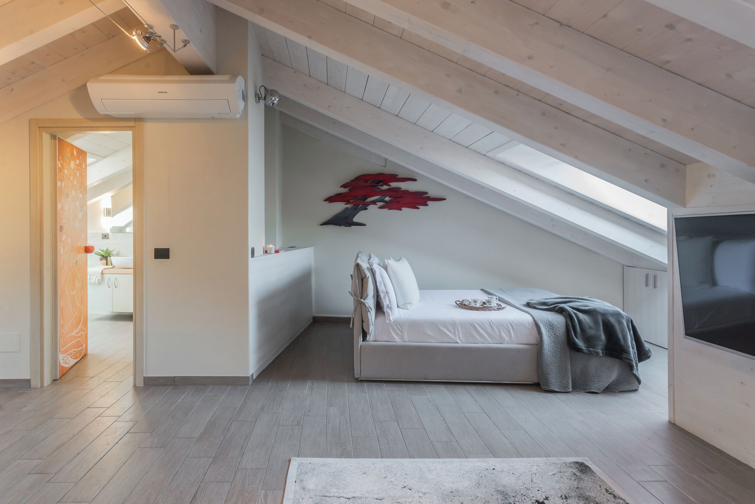 Ein modernes Schlafzimmer mit schrägen Wänden, einem Bett mit Decke, einem an der Wand montierten Fernseher und einem roten abstrakten Kunstwerk.