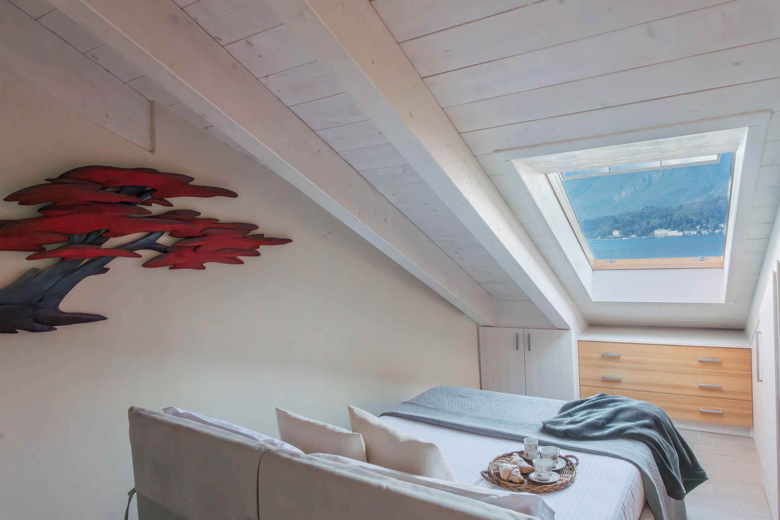 A bedroom with a skylight, a bed with a blanket, a bedside table with a cup and a basket, and a wall decoration.
