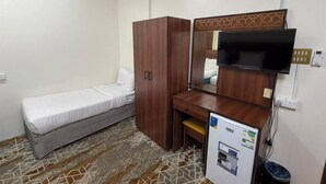A small room with a bed, a wooden wardrobe, a desk with a mirror, a television, and a small refrigerator. - MEHAD MADINAH QUBA (Madinah)