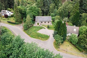 A stone building with a pitched roof, surrounded by dense greenery and a winding road. - Grade C Church in Cannich, Sleeps 2 w/ Parking (Beauly)