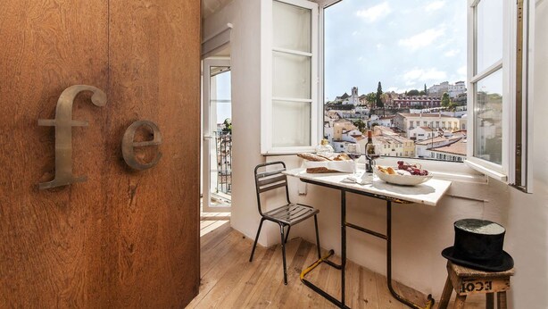 Een kamer met een houten vloer, een tafel met een fruitschaal en een wijnglas, een stoel en een raam met uitzicht op een stadsbeeld.