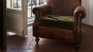 Een kamer met een bruine leren fauteuil, een groene bank, een vloerlamp en uitzicht door een open deur op een balkon met planten.
