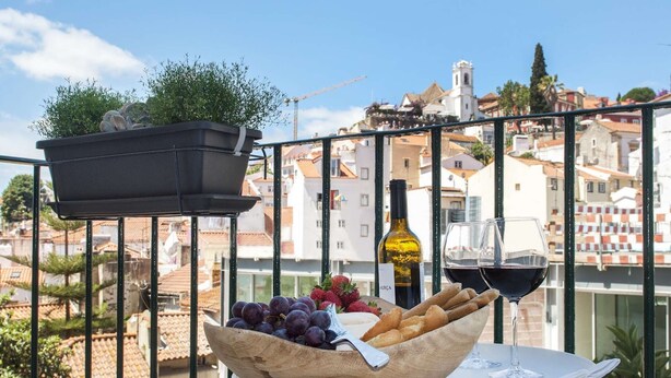 A balcony with a table set for a meal, featuring a bowl of grapes and bread, a bottle of wine, and two glasses of red wine. - Cheese&Wine™ | Alfama - Sto. Estevão Viewpoint 2 (Lisboa)