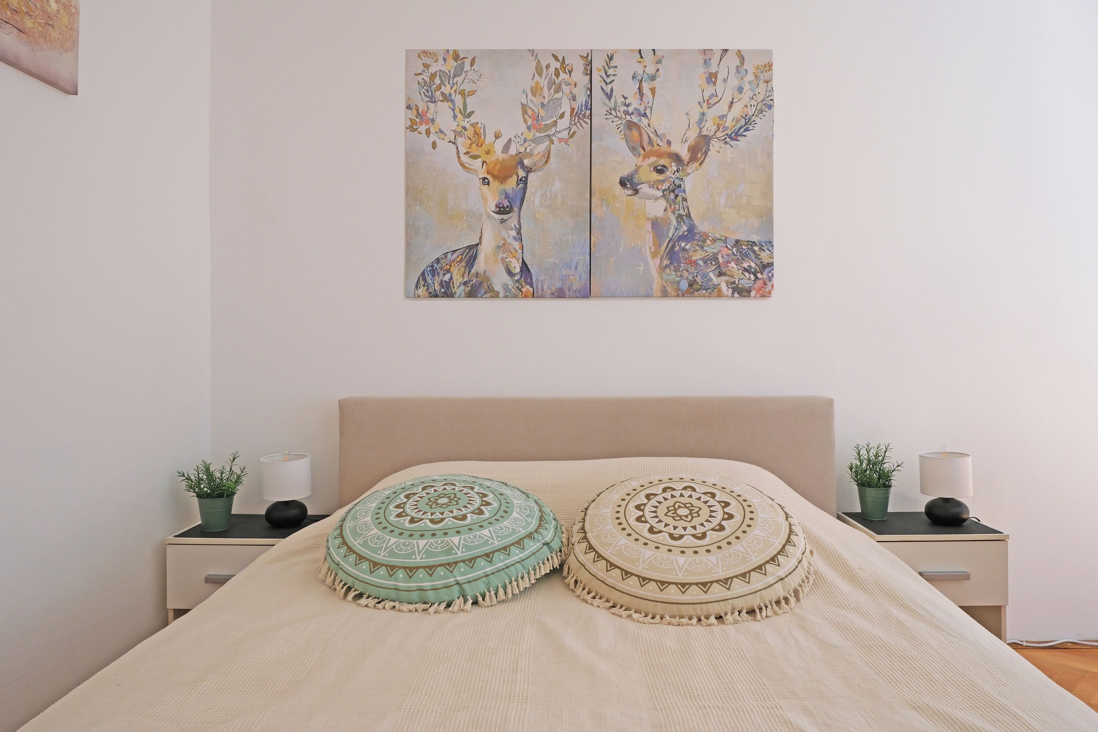 A bed with two decorative pillows, a nightstand with a plant and a lamp, and a painting of deer on the wall.