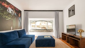 Ein Wohnzimmer mit einem blauen Sofa, einem kleinen Hocker, einem Fernseher auf einem Holzboden und einem großen Gemälde an der Wand.