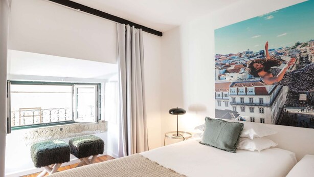 Ein Schlafzimmer mit einem großen Bett, einem Stadtbildgemälde, einem Fenster mit Vorhängen und einem kleinen Hocker.