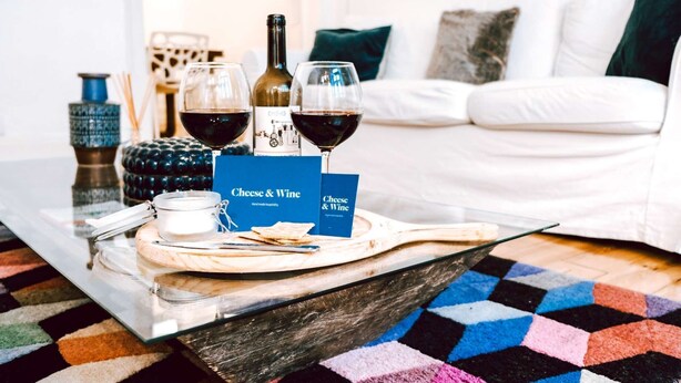 Una bandeja con queso y vino, una botella, copas y un menú sobre una mesa en una habitación con un sofá blanco y una alfombra estampada.