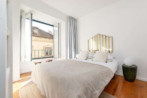 Ein Schlafzimmer mit einem großen Bett, Blick auf ein Gebäude durch ein offenes Fenster, einem Balkon und einem dekorativen Wandspiegel.
