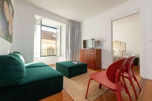 A living room with a green sofa, a red chair, a TV on a wooden stand, and a balcony with a view of buildings. - Cheese&Wine™ | Alfama - Sé Cathedral 2 (Lisboa)