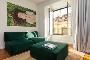 A room with a green sofa, a small ottoman, a painting on the wall, and a window with a view of buildings. - Cheese&Wine™ | Alfama - Sé Cathedral 2 (Lisboa)