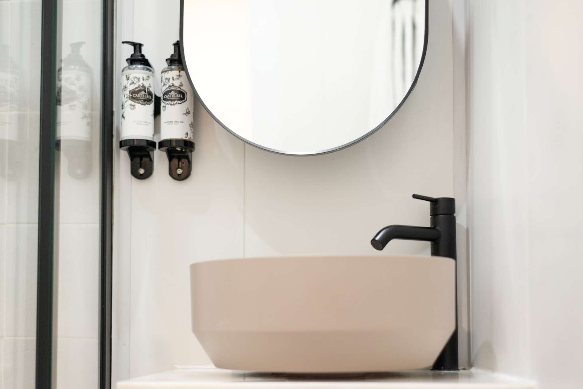 Een badkamer met een ronde spiegel, een beige wastafel en twee Castelfel flessen aan de muur.