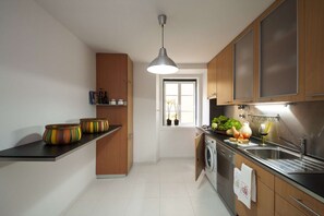 A modern kitchen with wooden cabinets, a black countertop, a hanging pendant light, and a window with plants. - Cheese&Wine™ | Chiado - Rossio Square 1 (Lisboa)