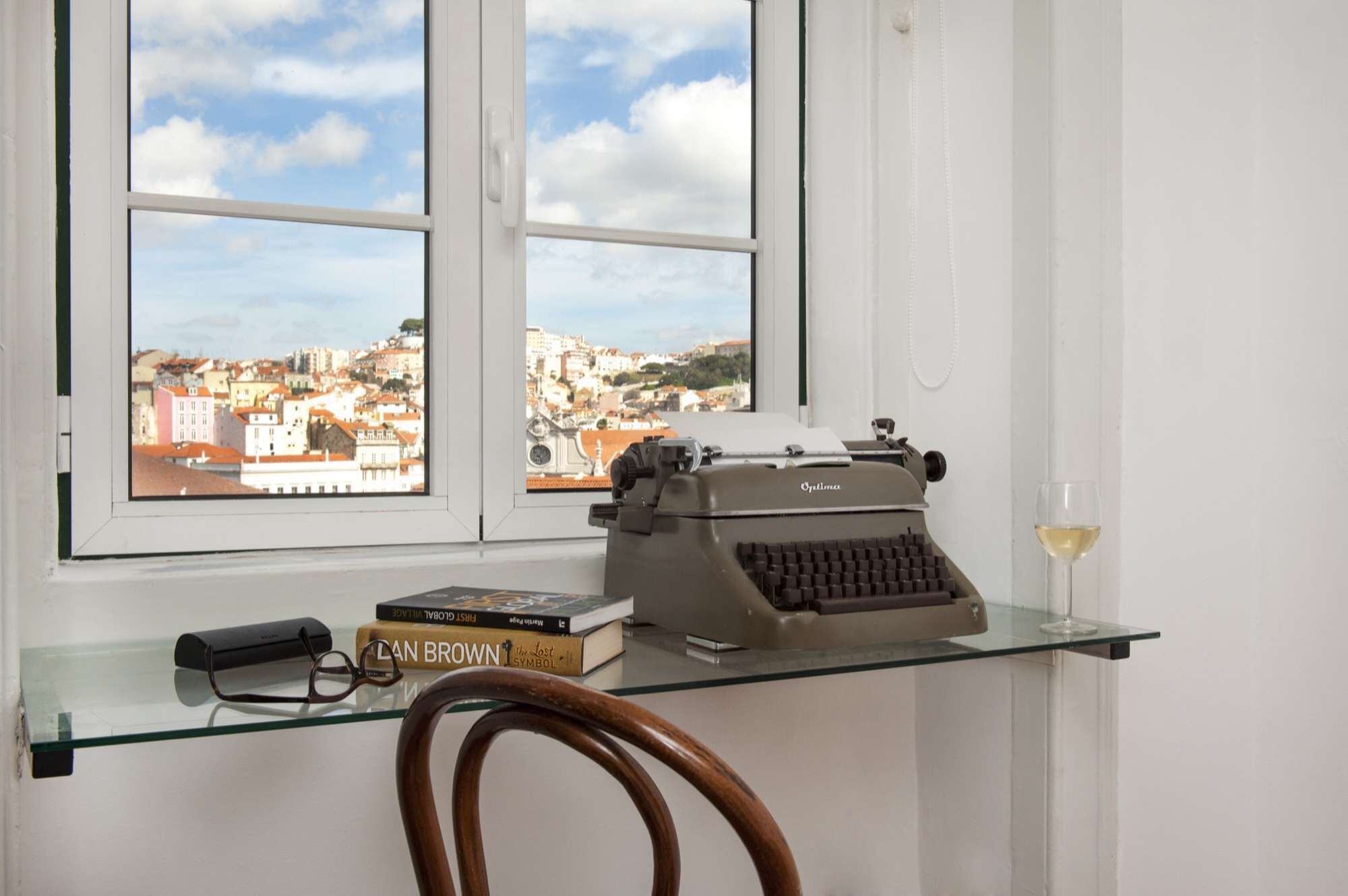 A room with a view of a cityscape through a window, a typewriter on a glass desk, a book titled 'The Lost Symbol,' and a wine glass.