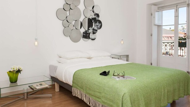 A bedroom with a bed, a green patterned bedspread, a wall art piece, a bedside table, and a window with a view. - Cheese&Wine™ | Chiado - Rossio Square 2 (Lisboa)