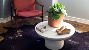 Ein Zimmer mit einem roten Sessel, einem runden weißen Tisch mit Pflanze und Buch sowie einem Kuhfellteppich.