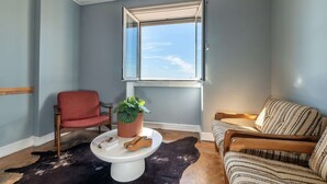 Ein Zimmer mit einem großen Fenster, einem roten Sessel, einem gestreiften Sofa, einem weißen Couchtisch, einer Topfpflanze und einem Lederteppich.