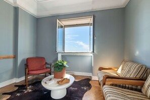 Ein Zimmer mit einem großen Fenster, einem roten Sessel, einem gestreiften Sofa, einem weißen Couchtisch, einer Topfpflanze und einem Lederteppich.