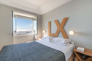 Ein Hotelzimmer mit einem Bett, einem großen Fenster mit Blick auf die Stadt, einem an der Wand befestigten Holzschild mit der Aufschrift 'LX' und Nachttischlampen.