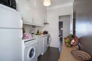 Eine kompakte Küche mit Waschmaschine, Backofen und einem Holztisch mit Früchten und einem Korbwägelchen.