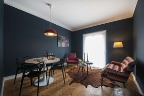 Ein Essbereich mit einem runden Tisch, Stühlen, einer roten Pendelleuchte, einem roten Sessel, einem gemusterten Sessel, einer Glasvase und einem Fellteppich.