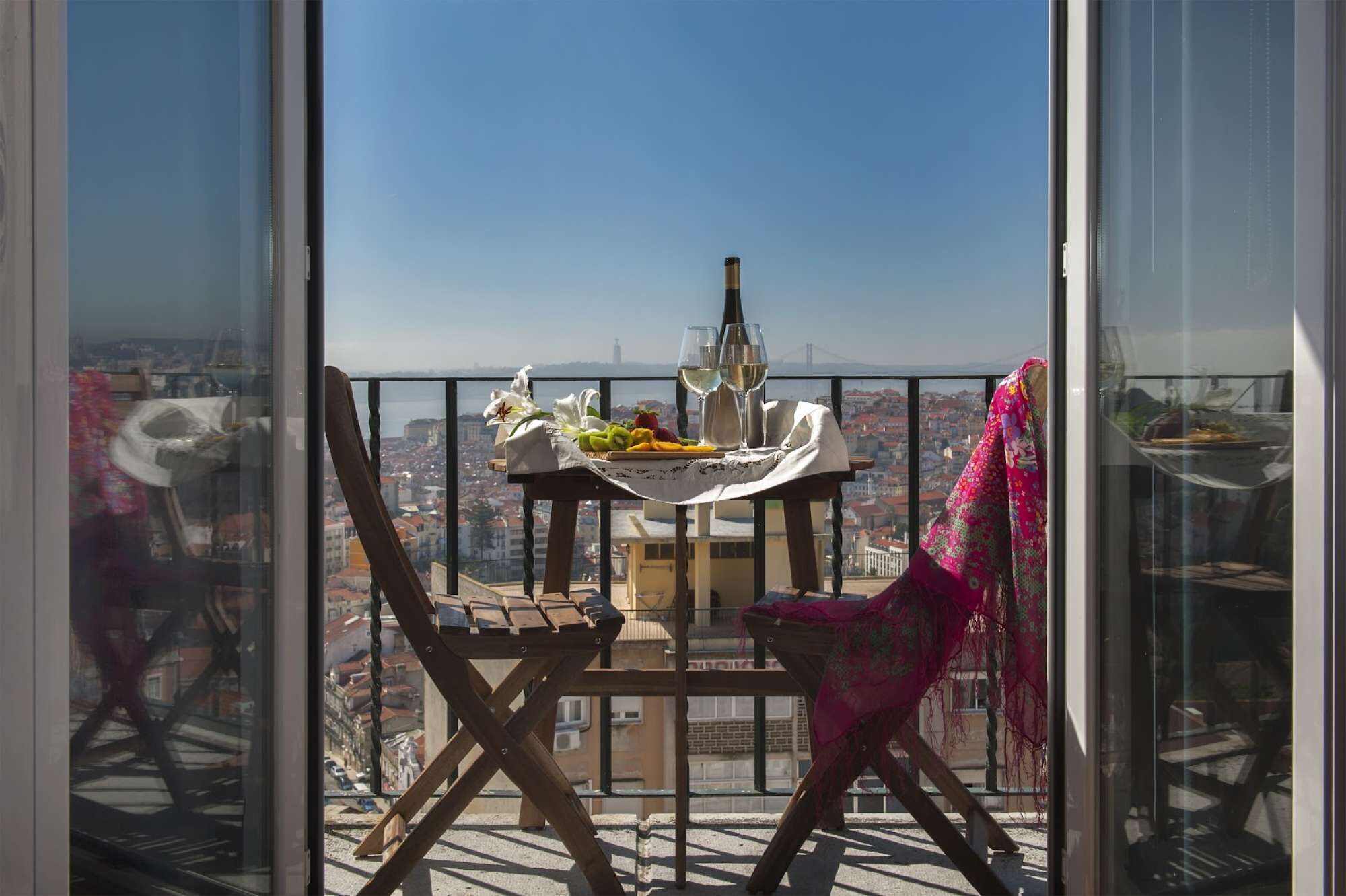 Un balcon avec une table dressée pour deux personnes, une bouteille et une chaise recouverte d’un tissu.