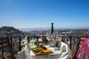 Ein Tisch mit einer Weinflasche, Gläsern und Obst auf einem Balkon mit Blick auf eine Stadtlandschaft.