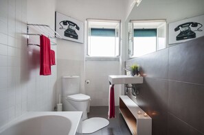 Ein modernes Badezimmer mit Toilette, einer Badewanne, einem Waschbecken mit Spiegel und zwei Fenstern.