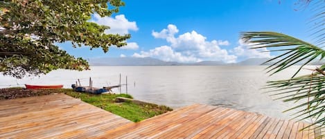 Un muelle de madera que da a un lago tranquilo con barcos, rodeado de follaje y un cielo despejado.