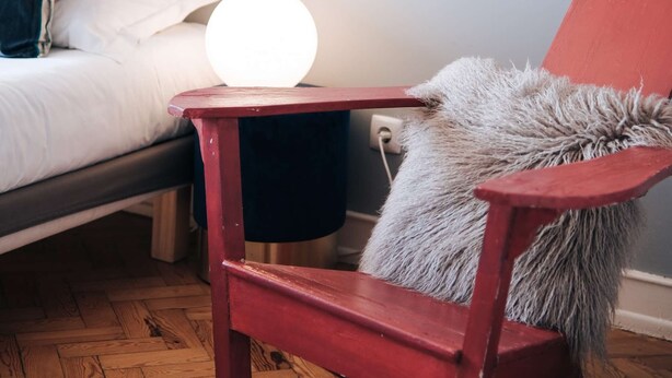 A room with a bed, a round table with a light, a chair with a fur throw, and a wall with a poster. - Cheese&Wine™ | Sra. do Monte - Viewpoint 3 (Lisboa)