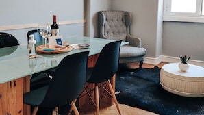 A modern room with a glass table, black chairs, a grey armchair, a round ottoman, and a window with natural light. - Cheese&Wine™ | Sra. do Monte - Viewpoint 3 (Lisboa)
