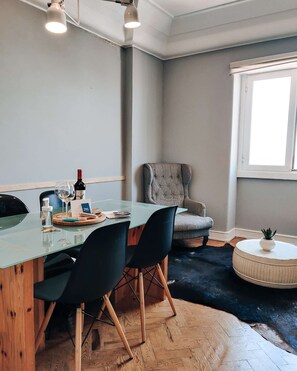 Ein modernes Zimmer mit Glastisch, schwarzen Stühlen, einem grauen Sessel, einem runden Pouf und einem Fenster mit Tageslicht.