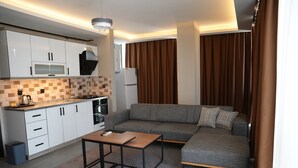 A modern kitchen with white cabinets, a microwave, and a stove. There's a gray sofa with a small table in front, and brown curtains on the windows. - Atlas Suit Otel (mersin)
