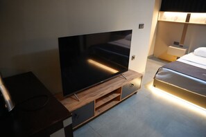A modern hotel room with a flat-screen TV on a wooden stand, a bed with a headboard, and a bedside table with a lamp. - Atlas Suit Otel (mersin)