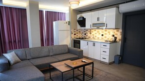 A compact living space with a grey sofa, a small wooden table, and a kitchenette with white cabinets and a microwave. - Atlas Suit Otel (mersin)
