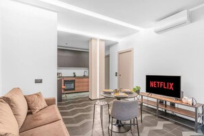 Ein modernes Wohnzimmer mit einer Couch, einem Esstisch mit Stühlen, einem Fernseher, der 'NETFLIX' zeigt, und einer Küche im Hintergrund.