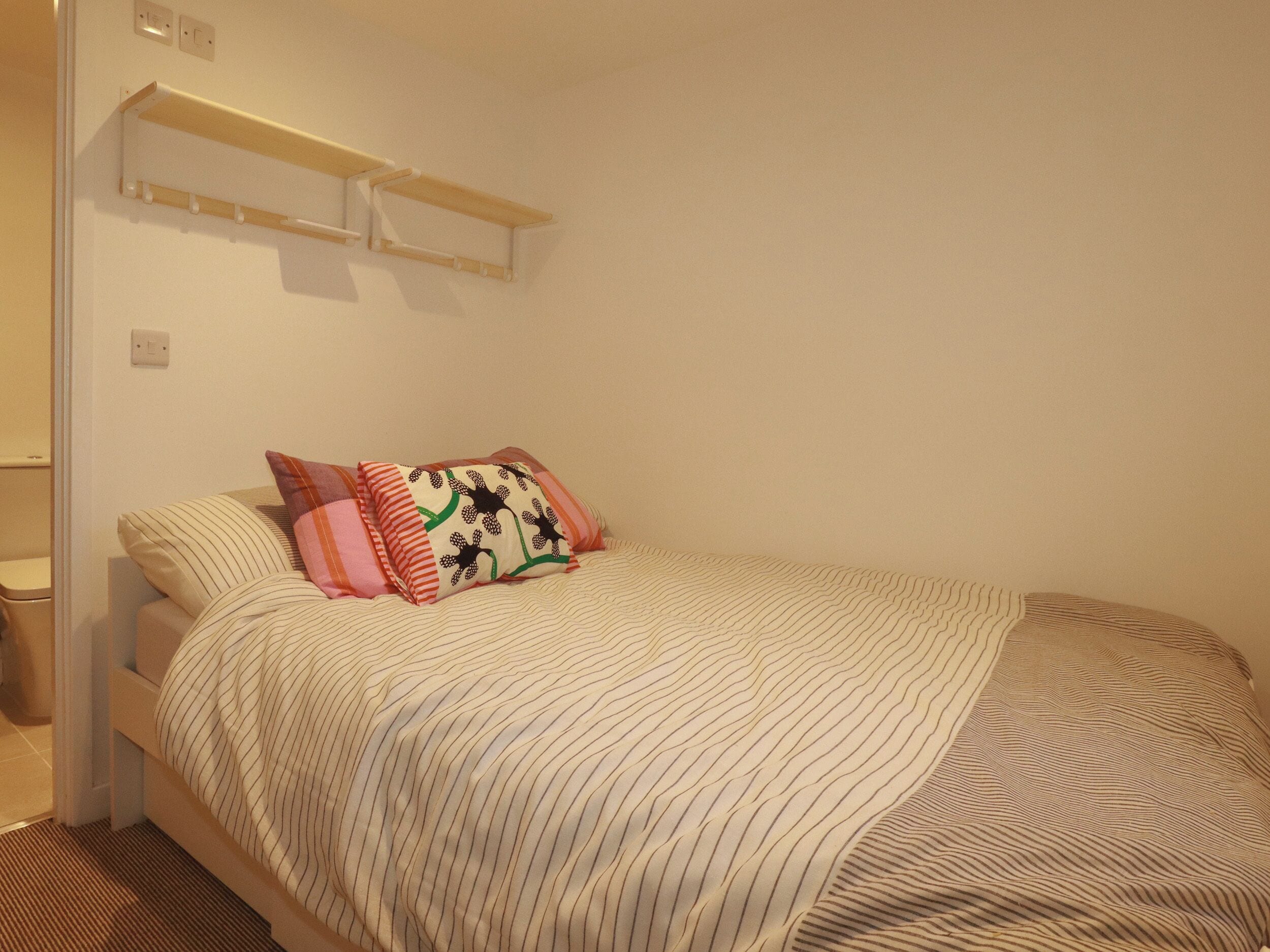 Een slaapkamer met een bed, kussens, een nachtkastje en een wandplank.