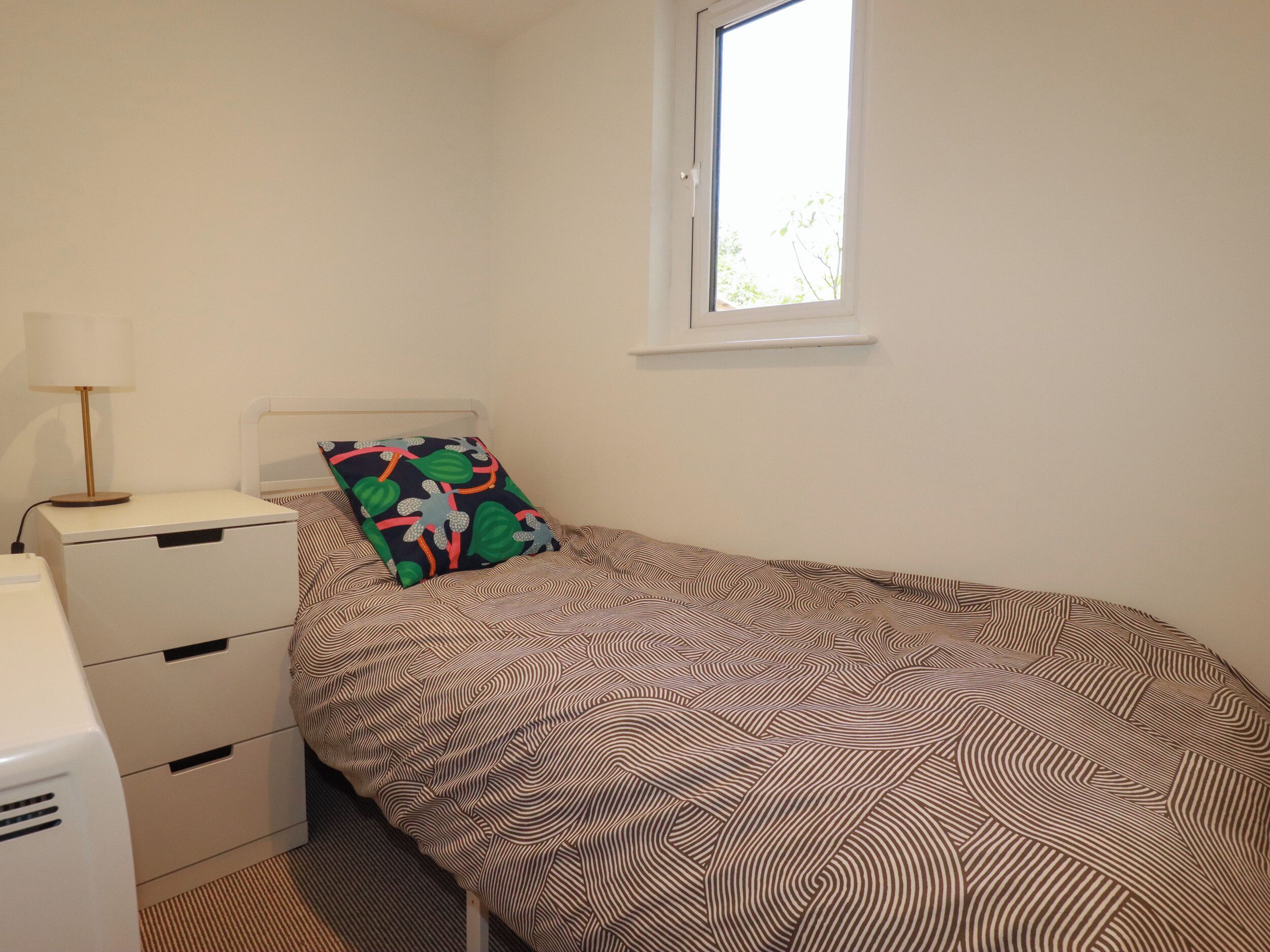 A small, single-bed room with a patterned bedspread, a bedside table with a lamp, and a window.